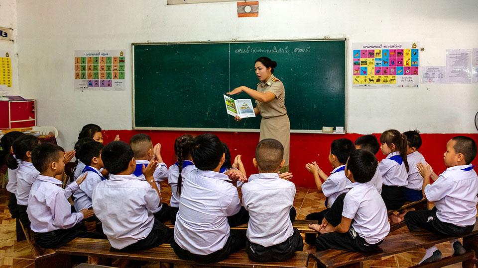 A female teacher stands in front of the class, telling them a story from the book she is holding